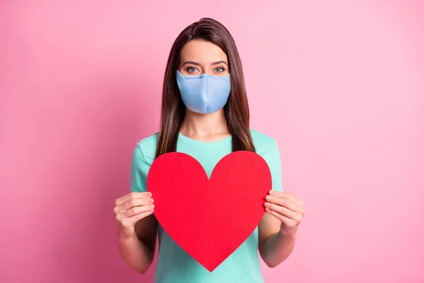 Foto retrato de menina bonita mantendo cartão em forma de coração vermelho vestindo máscara de tecido azul isolado no fundo de cor rosa pastel — Fotografia de Stock