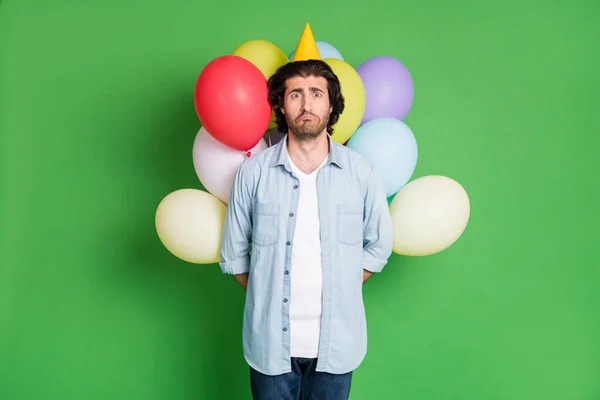 Foto de hombre mantenga globos detrás de la espalda mirada infeliz cámara usar papel cono gorra mezclilla jeans aislado color verde fondo — Foto de Stock