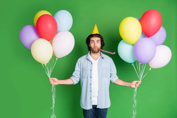Φωτογραφία του νεαρού αστείο άνδρα κατέχουν πολλά μπαλόνια σφυρίχτρα φορούν χάρτινο κώνο καπέλο τζιν πουκάμισο τζιν απομονωμένο πράσινο χρώμα φόντο — Φωτογραφία Αρχείου