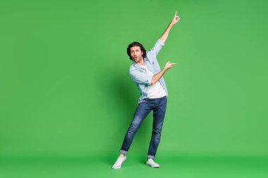 Full length photo of young stubbled man raise arms indicate fingers dance wear denim shirt jeans sneakers isolated green color background clipart