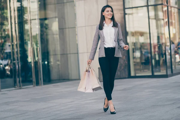 Полноразмерное фото счастливой симпатичной бизнесвумен, идущей по магазинам с сумками, прогуливающейся в блейзере по центру города — стоковое фото