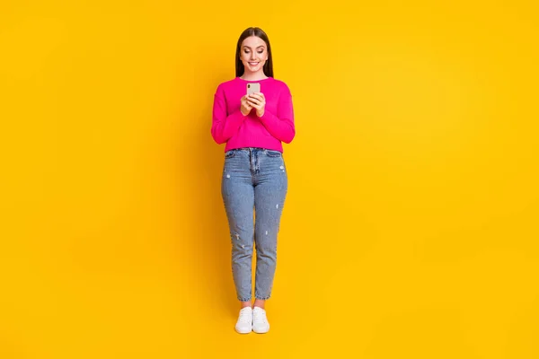 Full längd foto av härlig flicka hålla telefon look skärm bära rosa pullover jeans sneakers isolerad gul färg bakgrund — Stockfoto