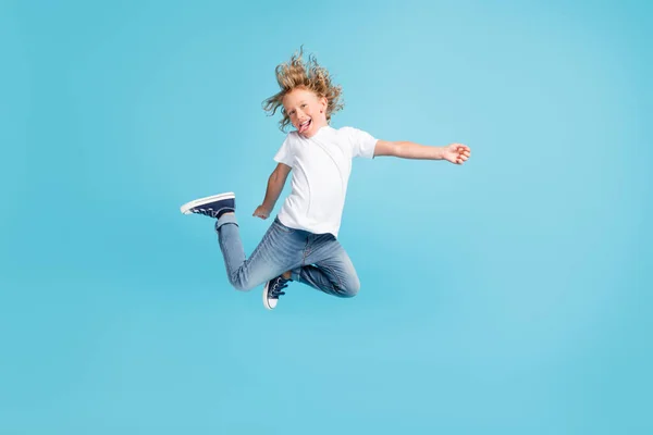 Фото маленького дитячого стрибка підняти ноги палицею на язику в білій сорочці джинси кросівки ізольовані синій колір фону — стокове фото