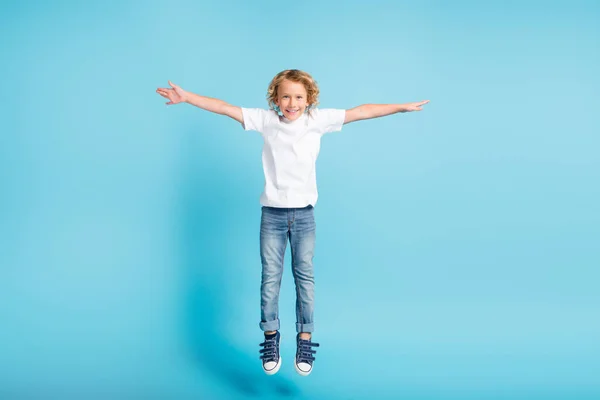 Фото маленького хлопчика, який стрибає підняти крила рук, одягнені в білі сорочки джинси кросівки ізольовані синій колір фону — стокове фото