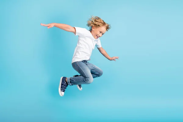 Фотографія профілю молодого хлопчика, що стрибає, рухаються руками, одягнені в білі сорочки джинси кросівки ізольовані синій колір фону — стокове фото