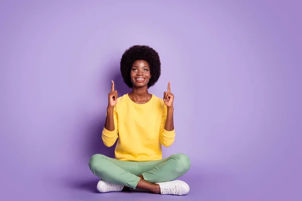Foto de longitud completa de afro chica americana sentarse piso piernas cruzadas dedo índice punto arriba espacio vacío aislado sobre fondo de color púrpura — Foto de Stock