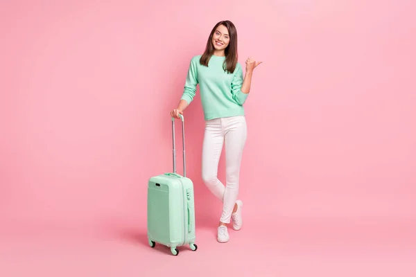 Photo d'une jolie jeune fille rêveuse pointant sur le copyspace avec un pouce tenant une valise isolée sur fond rose pastel — Photo