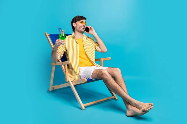 Amico, sono in paradiso. Full length foto uomo relax riposo resort sit chaise-lounge bere cocktail chiamare amico dire week-end indossare bianco giallo camicia pantaloncini a piedi nudi isolato colore blu sfondo — Foto Stock