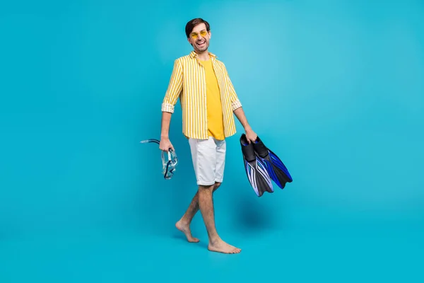 Full length foto positivo ragazzo turista andare a piedi spiaggia provare sport acquatici tenere maschera occhiali tubo pinne indossare giallo camicia a righe bianco corto piede nudo isolato blu colore sfondo — Foto Stock