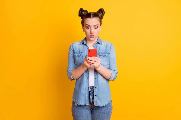 Фото божевільної шокованої дівчини-підлітка дві булочки дивляться телефон читати коментарі блог відкритий рот карантин повідомлення погані новини носити повсякденні джинсові сорочки ізольовані жовтий колір фону — стокове фото