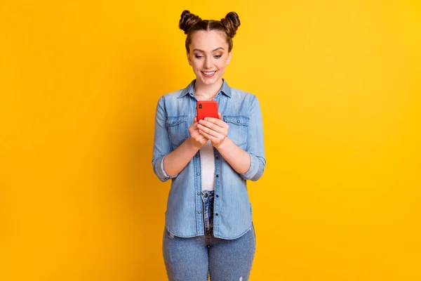 Фото чарівної смішної дівчини-підлітка дві булочки виглядають зацікавленими телефонними читаннями позитивні хороші коментарі блог абоненти носять повсякденну джинсову сорочку ізольований жовтий колір фону — стокове фото