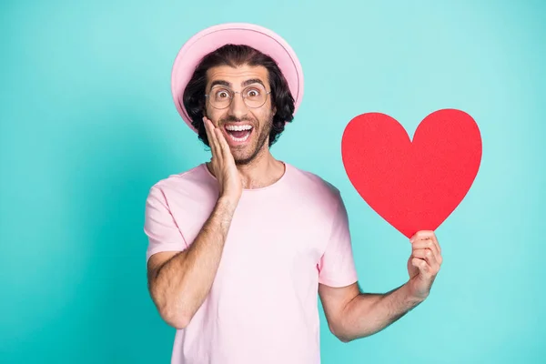 Retrato de tipo hipster optimista mano en la mejilla sostener papel corazón desgaste rosa gorra camiseta gafas aisladas sobre fondo de color verde azulado — Foto de Stock