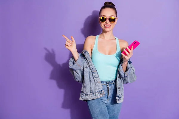 Positiv fröhlich Mädchen Student Blogger verwenden Smartphone zeigen soziale Netzwerk-Anzeigen Zeigefinger Kopierraum tragen Stil stilvolle trendige Jeans Jacke isoliert lila Farbe Hintergrund — Stockfoto