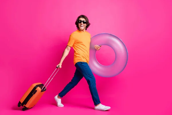 Foto de perfil de comprimento total do homem passo porão bagagem flutuante anel desgaste laranja t-shirt jeans sunglass calçado isolado cor-de-rosa fundo — Fotografia de Stock