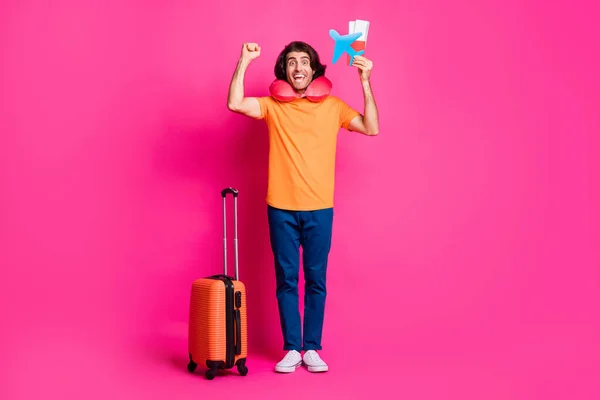 Foto de comprimento total do cara bilhetes de bagagem avião de papel levantar punho desgaste pescoço almofada laranja t-shirt jeans tênis isolado brilhante cor-de-rosa fundo — Fotografia de Stock