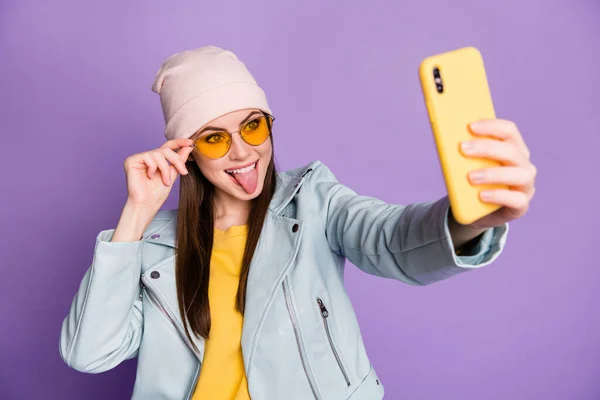 Фото стильної красивої молодої леді тримає телефон усміхненим липким язиком, щоб зробити селфі-підписників блогер носить сонцезахисні окуляри капелюх жовтий пуловер ізольований фіолетовий кольоровий фон — стокове фото