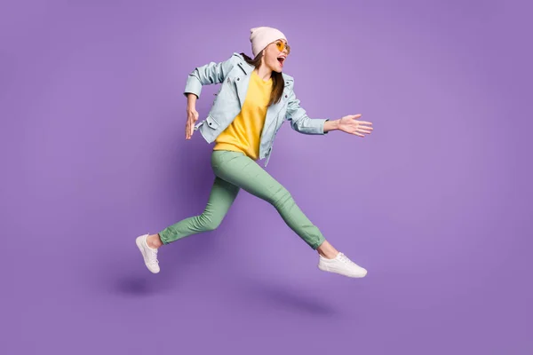 Full body profile side photo of surprised shocked cheerful girl jump run after discounts wear yellow green headwear pants isolated over violet color background