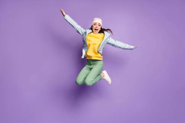 Volledige lengte foto van funky aantrekkelijke dame stijlvolle kleding springen hoog verheugende vliegende armen als vleugels vogel gek dragen casual hoed jas groene broek schoenen geïsoleerde paarse kleur achtergrond — Stockfoto