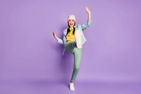 Full size photo of cool funky lady street clothes good mood dancing strange youngster moves raise leg high wear casual hat jacket pants shoes isolated purple color background
