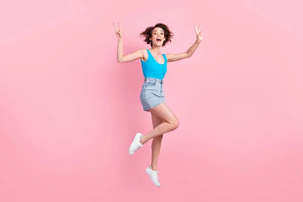 Full body size photo of pretty charming sporty young lady sorrindo open mouth jump raise hands showing v-sign wear denim mini skirt blue singlet sneakers isolated pink color background — Fotografia de Stock