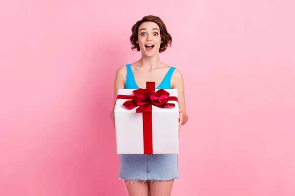 Photo of cute pretty young girl hold white surprise box red ribbon open mouth excited receive birthday gift celebrating wear denim mini skirt blue singlet isolated pink color background