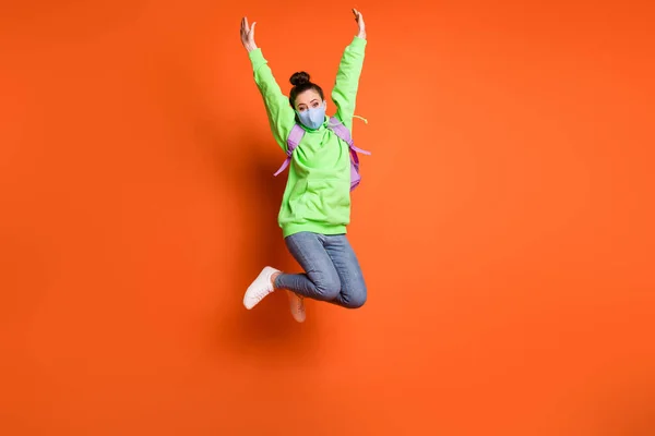 Retrato de foto de longitud completa de niña levantando las manos saltando usando máscara facial de tela azul aislada sobre fondo de color naranja vivo —  Fotos de Stock