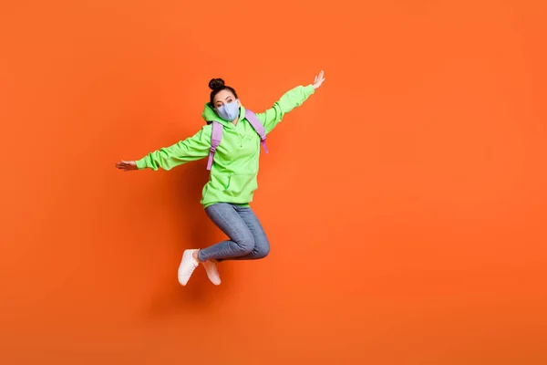 Full längd foto porträtt av skolflicka hoppa upp gör planet med händerna bär blå tyg ansikte mask isolerad på levande orange färgad bakgrund — Stockfoto