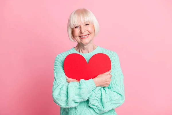 Фотопортрет милої бабусі, що обіймає червоне серце символ Валентина день ізольований на пастельному рожевому кольоровому фоні — стокове фото