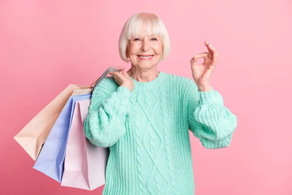 Фотопортрет бабушки, хранящей пакеты после покупки, показывающий хорошо улыбающийся знак, изолированный на пастельно-розовом фоне — стоковое фото