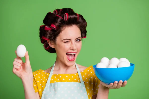 Foto de primer plano de linda encantadora chica joven rodillo peinado sonrisa parpadear ojo boca abierta sostener cuenco huevos presentando mostrando mejor uno usar delantal punteado camisa aislado color verde fondo —  Fotos de Stock