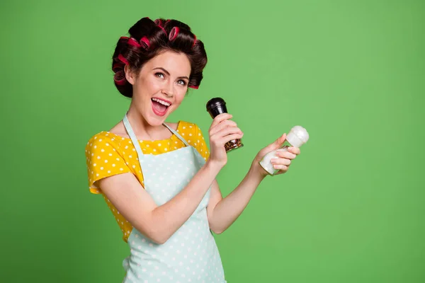 Foto von schönen charmanten funky junge Mädchen Rollenfrisur halten Pfefferstreuer Salzflasche offenen Mund vorgeben, mit Mikrofon singen tragen gepunktete Schürze Hemd isoliert grüne Farbe Hintergrund — Stockfoto