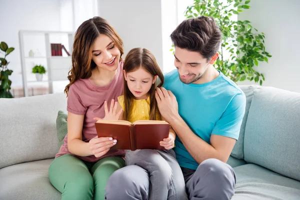 Foto van idyllische drie mensen zitten bank moeder vader kijken klein meisje kind lezen boek in huis binnen — Stockfoto