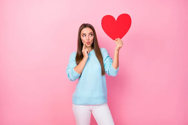 Bild av ganska listig dam hålla stora papper rött hjärta tänka på datum inbjudan svar skicka luft kyssar bära stickad blå tröja vita byxor isolerad pastell rosa färg bakgrund — Stockfoto