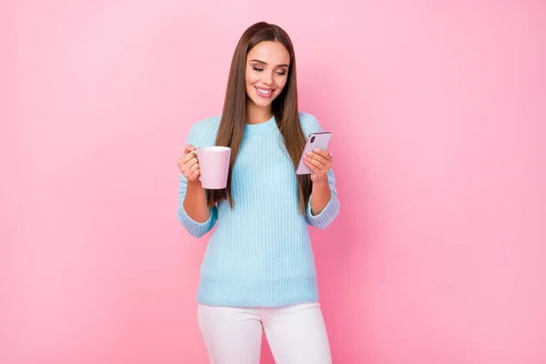Bild av attraktiv dam njuta av varm kaffe dryck hålla telefon kommunicera vänner pojkvän bära stickad blå pullover vita byxor isolerad pastell rosa färg bakgrund — Stockfoto