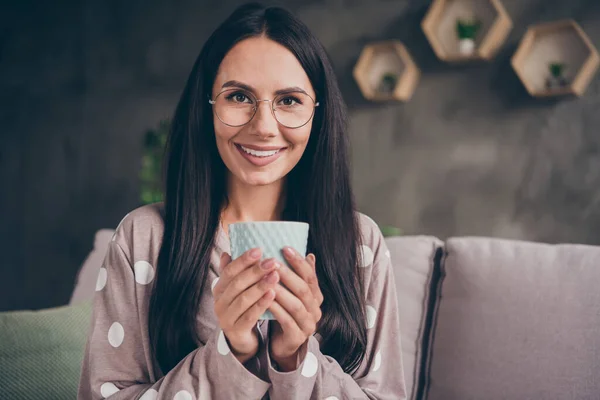 Portret van mooi optimistisch meisje houd beker dragen bril pijama thuis op bank — Stockfoto