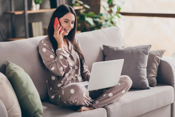 Фото милой оптимистической девушки сидеть писать ноутбук говорить телефон пижама носить дома — стоковое фото