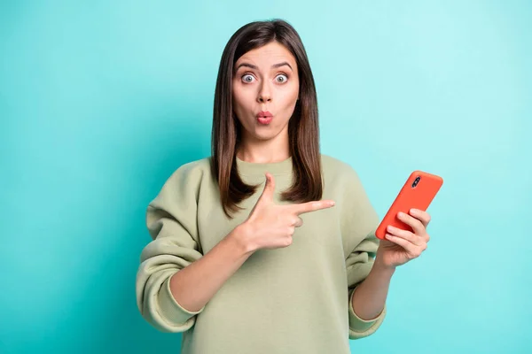 Foto portret van verbaasd vrouw wijzende vinger op het houden van telefoon in een hand geïsoleerd op levendige turquoise gekleurde achtergrond — Stockfoto