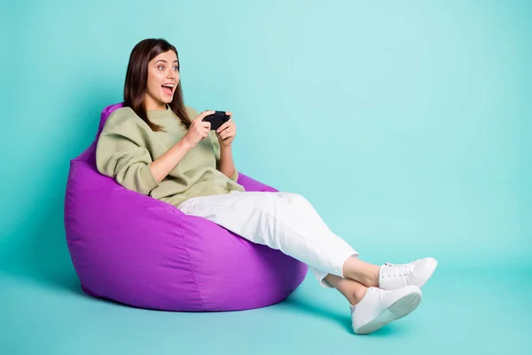 写真ポートレート興奮ゲーマーの女の子保持ジョイスティックのフルボディビューで2つの手に座っている紫色の豆の袋の椅子に隔離された鮮やかなターコイズ色の背景 — ストック写真