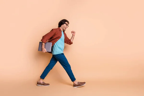Volledige lengte profiel foto van positieve man houden laptop lopen snelle slijtage bril bruin t-shirt geïsoleerde beige kleur achtergrond — Stockfoto