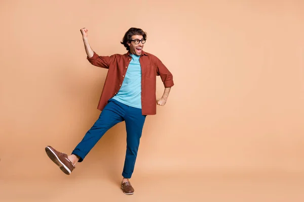 In voller Länge Foto von Kerl Tanz offenen Mund heben Faust Bein tragen Spezifikationen braun T-Shirt Hose Schuhe isoliert beige Farbe Hintergrund — Stockfoto