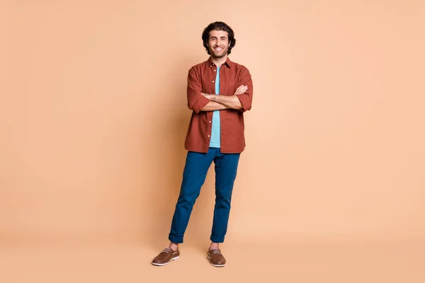 Pełna długość zdjęcie młodego faceta skrzyżowane ręce błyszczący uśmiech nosić brązowy t-shirt niebieskie spodnie buty odizolowany beżowy kolor tło — Zdjęcie stockowe