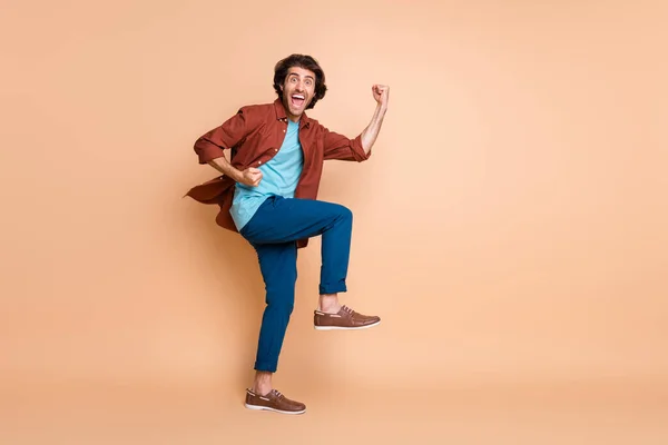 Foto de perfil de comprimento total do homem levantar punhos boca aberta olhar câmera desgaste marrom t-shirt calça azul sapatos isolado cor bege fundo — Fotografia de Stock