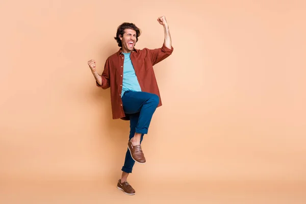 Фотографія повної довжини божевільного чоловіка, який піднімає кулаки відкритим ротом, носить коричневу футболку сині штани взуття ізольовані бежевий колір фону — стокове фото