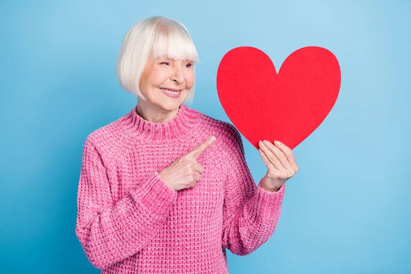 Fénykép portré idős nő keres mutató ujjal nagy piros szív kártya gazdaság egyik kezében elszigetelt pasztell kék színű háttér — Stock Fotó