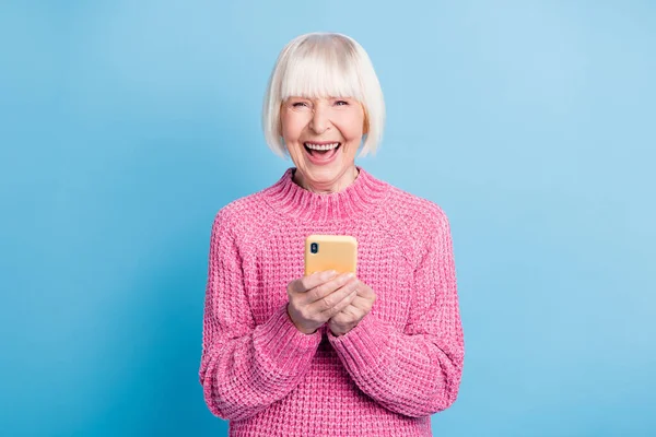 Photo portrait of excited old woman with open mouth holding phone in two hands isolated on pastel blue colored background