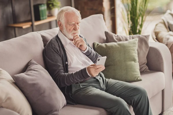 Oldalkép jóképű komoly öregember öltözött fehér ing kezében modern eszköz ül kanapé beltéri lakás — Stock Fotó