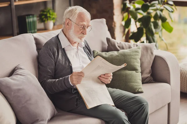 Foto de atento anciano vestido de blanco camisa gafas sentado sofá lectura últimas noticias dentro de casa plana interior casa —  Fotos de Stock