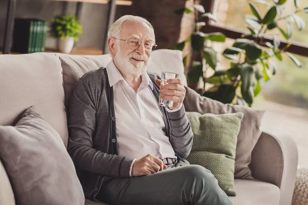 Foto de hombre viejo positivo vestido camisa blanca gafas sentado sofá que descansa agua potable en el interior interior casa plana — Foto de Stock