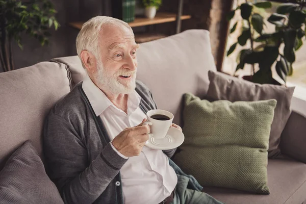Fotografie legrační důchodce nosit šedý svetr sedací gauč s odpočinkem pití kávy vnitřní dům byt — Stock fotografie