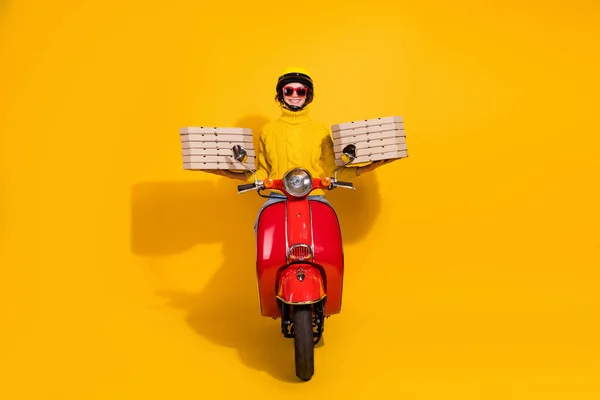 Retrato fotográfico de una repartidora sosteniendo pilas de cajas de pizza en dos manos montando una moto retro roja aislada sobre un fondo de color amarillo vivo — Foto de Stock